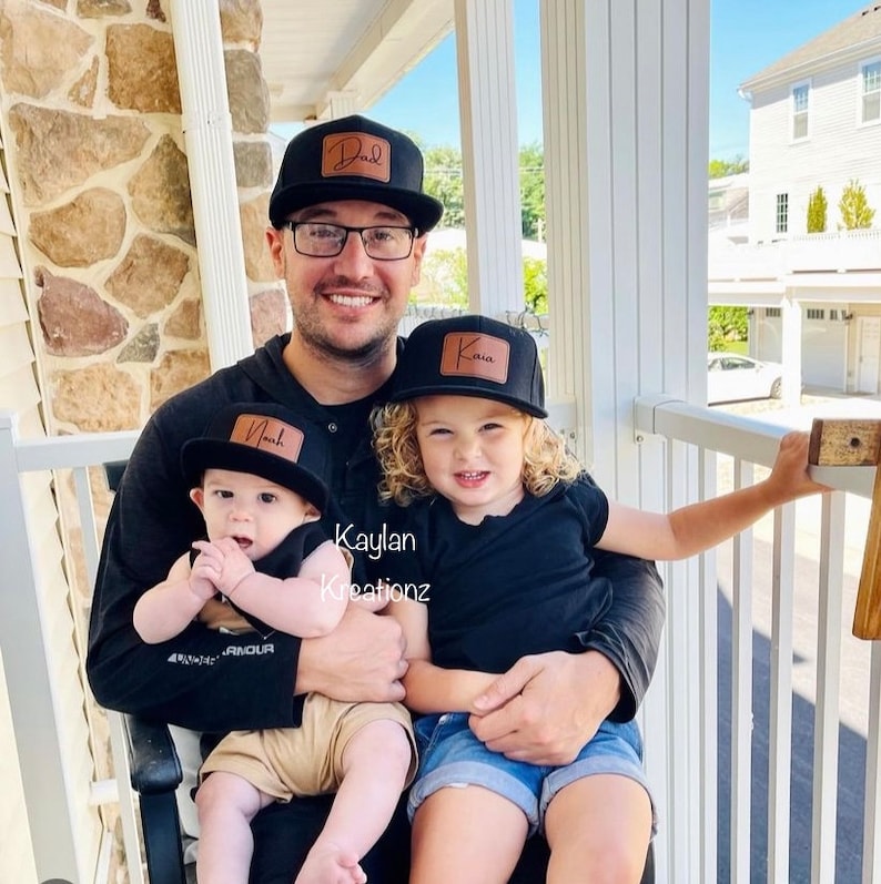 May include: A father and his two children are wearing black baseball caps with personalized leather patches. The father's cap says "Dad", the older child's cap says "Kava", and the younger child's cap says "Noah". The father is holding both children in his arms. The text "Kaylan Kreations" is visible in the image.