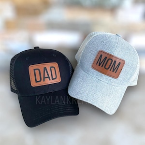 May include: Two black and gray mesh trucker hats with brown leather patches. The patch on the black hat says "DAD" and the patch on the gray hat says "MOM".