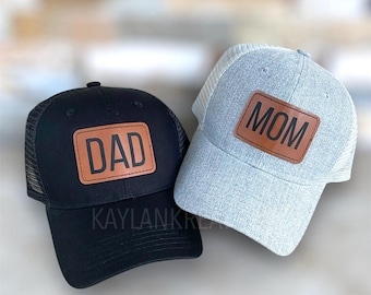 May include: Two black and gray mesh trucker hats with brown leather patches. The patch on the black hat says "DAD" and the patch on the gray hat says "MOM".