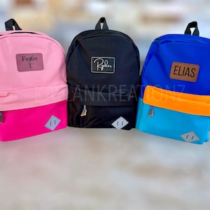 May include: Three personalized backpacks in pink, black, and blue. Each backpack has a contrasting bottom panel and a patch with a name: Kaylee, Ryder, and Elias. The backpacks have a diamond-shaped accent.