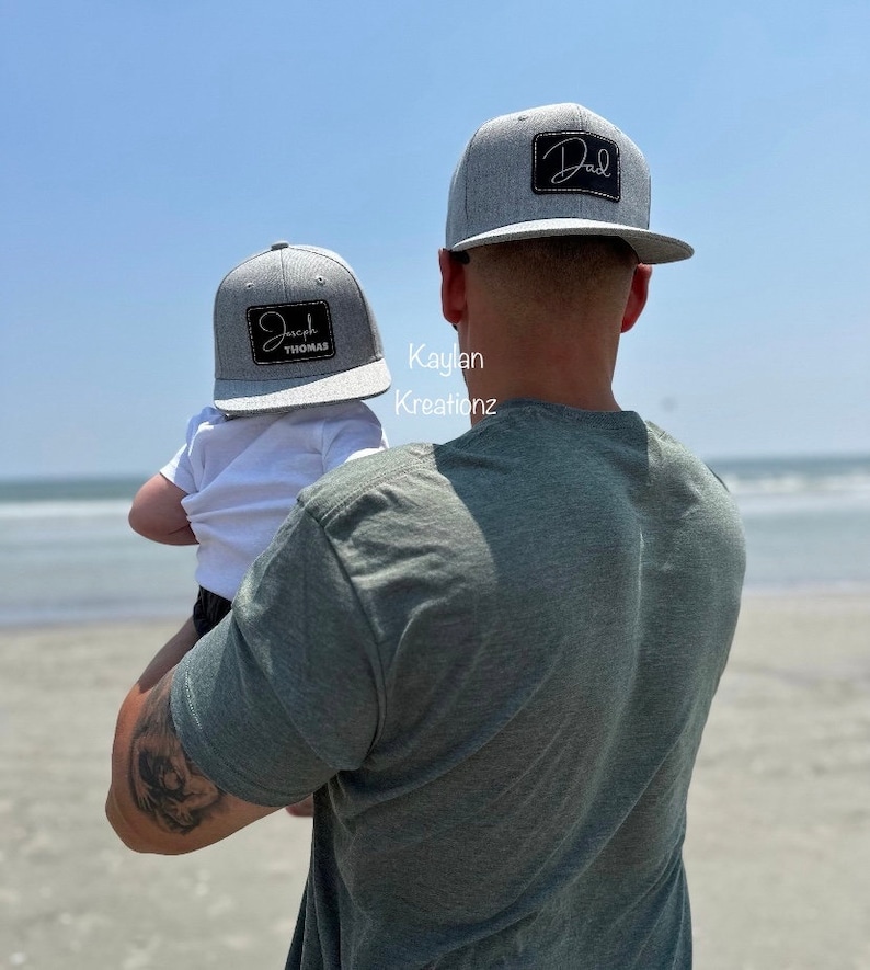 May include: A father and son wearing matching gray baseball caps with black embroidered text. The father's cap says "Dad" and the son's cap says "Joseph Thomas". The father is holding the son in his arms. They are standing on a beach.