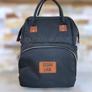 May include: Black diaper bag backpack with brown leather accents and a brown leather patch with the text "JOSIAH LIAM" in black.
