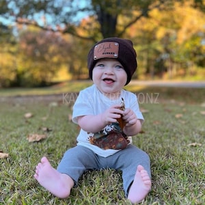 May include: A brown knit beanie with a leather patch that says "Elijah Gunner" on it. The beanie is worn by a young child sitting on the grass.
