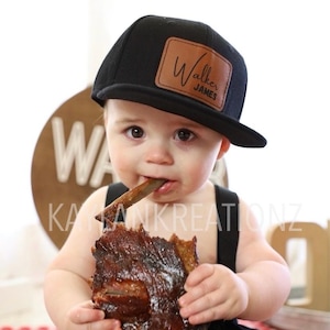 May include: A baby wearing a black baseball cap with a brown leather patch that says "Walker James" is eating a large piece of barbecue ribs.