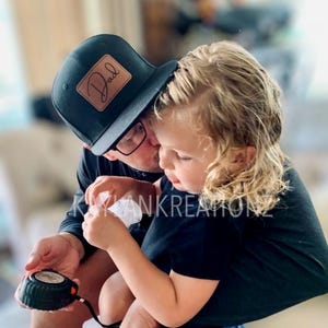 May include: A man wearing a black baseball cap with a brown leather patch that says "Dad" is holding a young child. The child is wearing a black t-shirt and blue jeans.