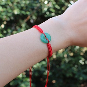 Puede incluir: Un brazalete de cuerda roja con un dije de jade verde. El dije es un disco redondo con un agujero en el centro.