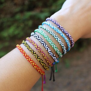Peut inclure: Un ensemble de huit bracelets d'amitié colorés avec un motif tressé. Les bracelets sont faits d'un cordon fin et présentent une variété de couleurs, notamment le rouge, le jaune, le noir, le vert, le bleu et le violet.