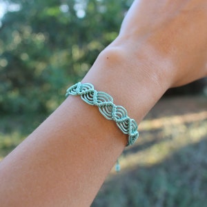 May include: A light blue friendship bracelet with a woven pattern. The bracelet is on a person's wrist.