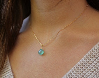 May include: A delicate gold necklace featuring a small, teardrop-shaped, light blue pendant. The pendant hangs from a fine gold chain, creating a simple and elegant design. The necklace is shown on a person wearing a beige sweater.