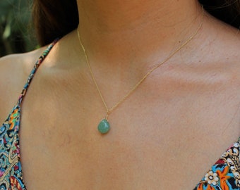May include: A gold chain necklace with a light green teardrop-shaped pendant. The necklace is worn by a person with a patterned top.