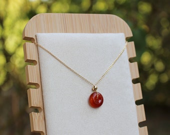 May include: A gold chain necklace with a round, red gemstone pendant displayed on a wooden stand.