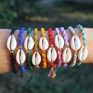 Può includere: Un set di 12 braccialetti colorati con conchiglie bianche e perline dorate. I braccialetti sono realizzati con corda intrecciata e sono disponibili in una varietà di colori, tra cui nero, blu, verde, giallo, rosso, rosa e viola.