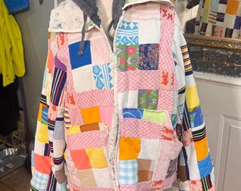 May include: A patchwork jacket with a gray hood and zipper closure. The jacket is made of various fabric squares in colors like pink, blue, yellow, and green. It is displayed on a wooden mannequin stand.