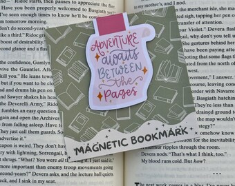 May include: A pink and white magnetic bookmark with the text "Adventure Awaits Between the Pages" in a whimsical font. The bookmark is placed on an open book, with a patterned background.