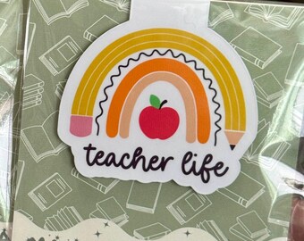 May include: A magnetic bookmark featuring a rainbow design with a red apple in the center. The bookmark has the words "teacher life" and "MAGNETIC BOOKMARK" printed on it. The background has a pattern of green books.
