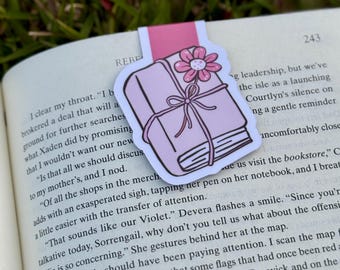 May include: A pink and white bookmark featuring a stack of books tied with a bow and topped with a flower. The bookmark is inserted into a book with text visible on the pages, set against a backdrop of green grass.