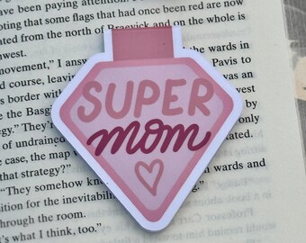 May include: A pink and white bookmark with the words "SUPER MOM" in a script font and a heart symbol. The bookmark is diamond-shaped and is placed on the pages of a book.