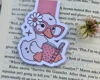 May include: A whimsical bookmark featuring a cartoon duck wearing glasses and reading a book. The duck has a daisy on its head and is set against a pink bookmark tab. The book has pink polka dots.