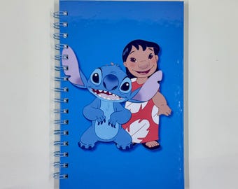 May include: A blue spiral-bound notebook featuring a colorful illustration of the Disney characters Stitch and Lilo. Stitch is blue with large ears, and Lilo is wearing a red dress with white floral patterns. The notebook has a glossy cover.