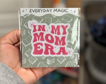 May include: A sticker with the text "IN MY MOM ERA" in pink, retro-style lettering. The sticker is on a background of green books and the words "EVERYDAY MAGIC" are at the top. The sticker is in a clear plastic sleeve.