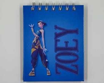 May include: A blue spiral-bound notepad featuring a stylized illustration of a character in a blue and yellow outfit. The word "ZOEY" is printed vertically in large letters on the right side of the notepad.