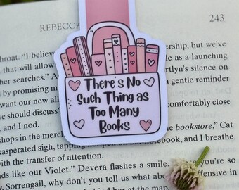 May include: A pink and white bookmark with the text "There's No Such Thing as Too Many Books" and illustrations of books and hearts. The bookmark is placed on an open book with text.