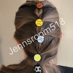 May include: Five colorful hair ties with different sports ball designs. The hair ties are in a ponytail on a person's head. The hair ties are black, white, orange, yellow, and green.