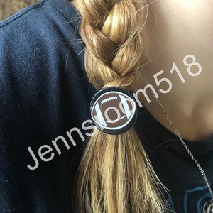 May include: A black and white football design button is used as a hair tie on a blonde braid.