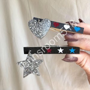 May include: Two decorative clothespins. One features a silver glitter heart, the other a silver glitter star. The black clothespins have red, white, and blue heart and star accents. These decorative pins are ideal for crafts and home decor.