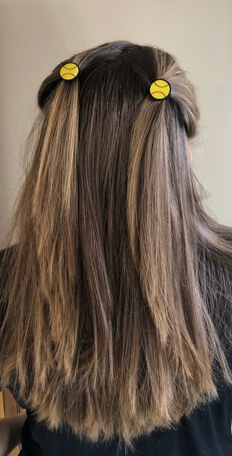 May include: A person with long brown hair with blonde highlights wearing two black hair ties with yellow softball designs.