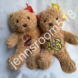 May include: Two brown teddy bears wearing baseball-themed outfits. One bear is wearing a red and white baseball outfit with the number "2" on it. The other bear is wearing a yellow and white baseball outfit with the number "13" on it.