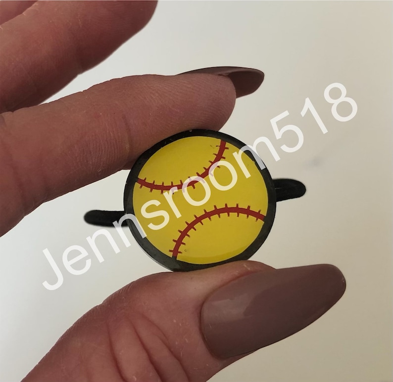 May include: A black and yellow softball charm with red stitching. The charm is attached to a black elastic hair tie.