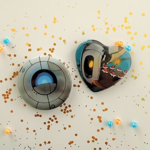 Può includere: Due bottoni rotondi con spilla con sfondo bianco e contorni neri. Il bottone a sinistra presenta un robot grigio con un occhio blu. Il bottone a destra ha la forma di un cuore e presenta un robot che tiene una torta di compleanno con candele.