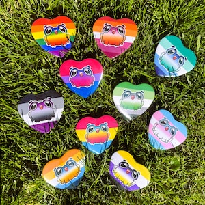 May include: A collection of heart-shaped pins featuring a cartoon frog with glasses. Each pin displays a different LGBTQ+ pride flag design, including rainbow, lesbian, bisexual, asexual, pansexual, non-binary, transgender, and aromantic flags.