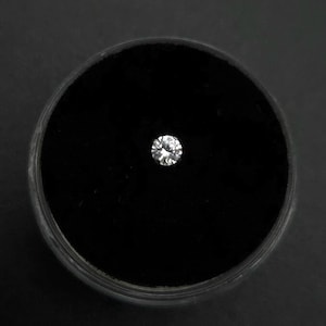 May include: A small, round, brilliant-cut diamond set against a black velvet background within a circular container. The diamond sparkles with multiple facets, reflecting light. The container is set against a dark, matte surface.