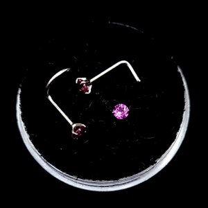 May include: A set of three nose studs and a nose ring, displayed in a clear container against a black background. The studs have a silver-colored post with a small, round, dark pink gemstone. The nose ring has a small, round, bright pink gemstone.