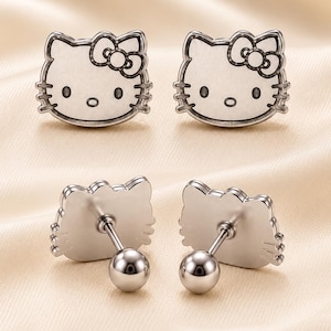 May include: A pair of silver-toned stud earrings shaped like the face of a cat character with a bow on one side. The front features engraved facial details including eyes, nose, and whiskers. The back shows a post with a round ball-shaped backing.
