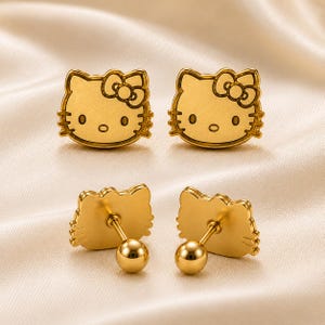 May include: A pair of gold-toned stud earrings shaped like the face of a cat character with a bow on one ear. The front shows the engraved facial features, while the back displays the post and a spherical screw-on backing for secure attachment.