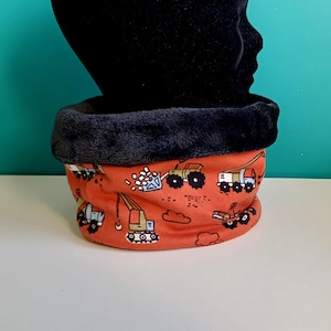 Peut inclure: Un tour de cou orange avec une doublure en peluche noire. Le tissu orange présente un motif répété de véhicules de chantier, notamment des camions, des excavatrices et des bulldozers. Le tour de cou est conçu pour garder le cou au chaud.