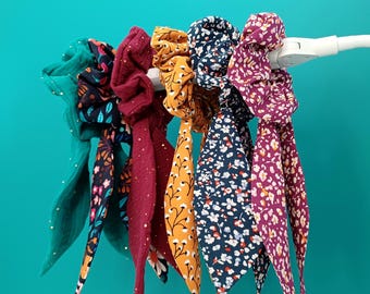 Puede incluir: Una colección de coleteros coloridos con largas colas de tela. Los coleteros presentan varios estampados, incluyendo estampados florales y lunares, en tonos turquesa, burdeos, amarillo mostaza y azul marino. Se muestran sobre un fondo turquesa.