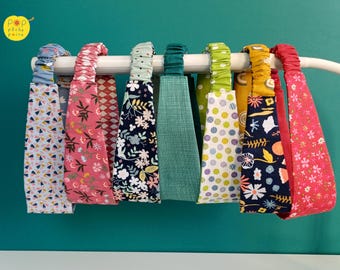 Puede incluir: Una colección de siete diademas de tela exhibidas en una barra blanca. Las diademas presentan varios patrones, incluyendo estampados florales, lunares y verde sólido, con secciones traseras elásticas para un ajuste flexible.