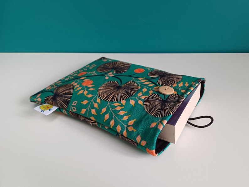 Puede incluir: Una funda de libro verde con un patr&oacute;n floral en tonos naranja, negro y blanco. La funda tiene un cierre de bot&oacute;n y una correa negra.