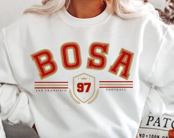 Può includere: Felpa bianca con scritta rossa e dorata "BOSA" e uno scudo sotto che dice "SAN FRANCISCO 97 FOOTBALL".