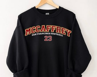Può includere: Felpa nera con una grafica ricamata in oro e rosso che dice "McCaffrey" con "San Francisco Football" e il numero "23" sotto.