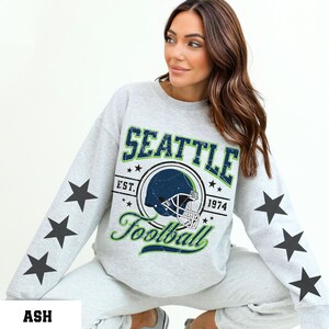 May include: Light gray sweatshirt with the text "SEATTLE Football 1974" and a football helmet graphic. Black star graphics are printed on the sleeves. The sweatshirt is made of a soft material.