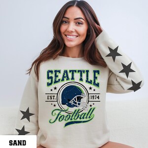 May include: Sand-colored sweatshirt with a Seattle Football graphic. The design includes a football helmet, the word "SEATTLE", and the text "EST. 1974". The sleeves feature black star graphics.