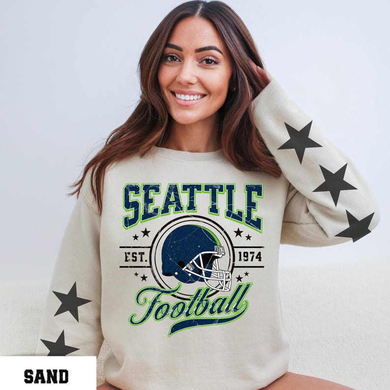 May include: Sand-colored sweatshirt with a Seattle Football graphic. The design includes a football helmet, the word "SEATTLE", and the text "EST. 1974". The sleeves feature black star graphics.