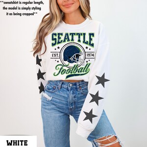 May include: White sweatshirt with a Seattle Football graphic. The design includes a football helmet inside a circle, the text "SEATTLE", "EST. 1974", and "Football". Black star graphics are on the sleeves.