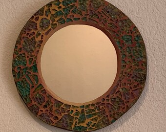 May include: A round mirror with a mosaic frame. The frame is made of small, irregular pieces of ceramic in shades of brown, green, and yellow.