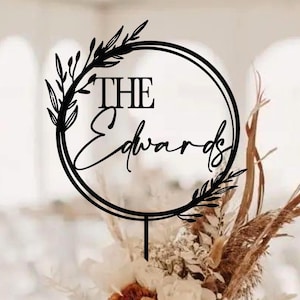 May include: A black cake topper with the words "THE Edwards" in a circular design, accented with leaf and floral details. The topper is placed on a cake decorated with white frosting and flowers, including a prominent orange rose.